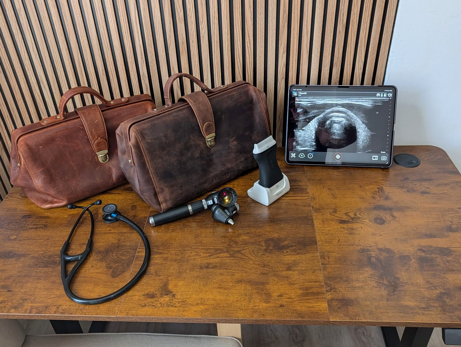 Twee leren dokterstassen, een stethoscoop, een otoscoop en een tablet met een echobeeld op een houten tafel